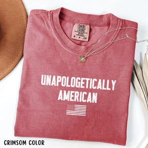 Może przedstawiać: Karmazynowa koszulka z napisem "UNAPOLOGETICALLY AMERICAN" i małą grafiką amerykańskiej flagi. Koszulka ma okrągły dekolt i kr&oacute;tkie rękawy. Złoty naszyjnik z wisiorkiem jest zarzucony na koszulkę.
