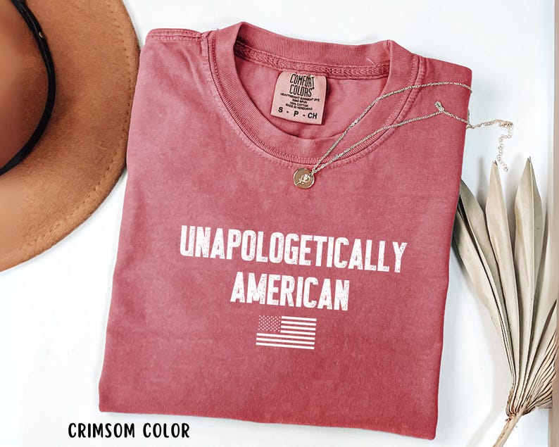 Może przedstawiać: Karmazynowa koszulka z napisem "UNAPOLOGETICALLY AMERICAN" i małą grafiką amerykańskiej flagi. Koszulka ma okrągły dekolt i kr&oacute;tkie rękawy. Złoty naszyjnik z wisiorkiem jest zarzucony na koszulkę.