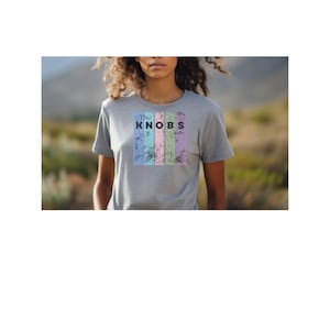 May include: A gray t-shirt with the text "The KNOBS" in black letters, set against a backdrop of vertical pastel stripes. The shirt is short-sleeved and appears to be made of a soft material.
