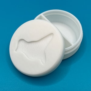 Pode incluir: Caixa de comprimidos de plástico branco com um compartimento dividido. A tampa tem um desenho em relevo de uma silhueta de animal estilizada.
