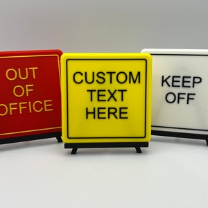 May include: Three small, square signs with black text on different coloured backgrounds. The first sign is red with yellow text that says "OUT OF OFFICE". The second sign is yellow with black text that says "CUSTOM TEXT HERE". The third sign is white with black text that says "KEEP OFF".