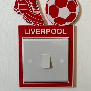 May include: Red and white light switch cover with a football and a football boot on top. The cover has the word "LIVERPOOL" printed on it.