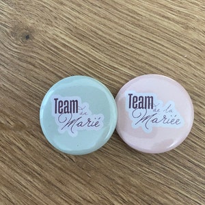 Peut inclure: Deux boutons ronds avec le texte "Team de la Mariée" et "Team de Marié" en écriture cursive. Le bouton de gauche est bleu clair et le bouton de droite est rose clair.