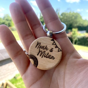 Peut inclure: Porte-clés en bois avec une étiquette ronde de couleur brun clair. L'étiquette porte le texte "Noah & Milan" gravé, ainsi qu'un arc-en-ciel et une guirlande de drapeaux.