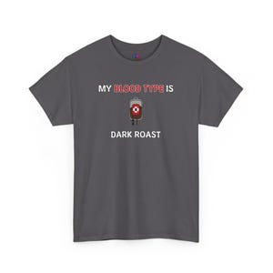Puede incluir: Camiseta gris oscuro con el texto "My blood type is dark roast" y una imagen de dibujos animados de una bolsa de sangre con café dentro.