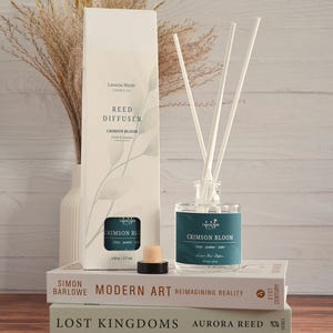 May include: A reed diffuser set with a clear glass bottle, white reeds, and a matching box. The box and bottle feature teal labels with the text "Crimson Bloom". The set is displayed on top of books with decorative dried plants in a white vase.
