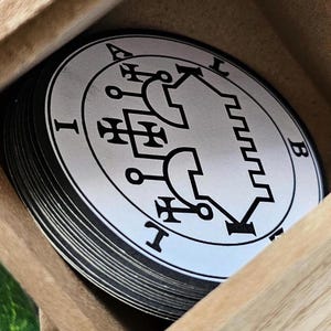 May include: A wooden box with a lid open, revealing a stack of round, white cards with black sigils. The sigil on the top card is a circle with a complex design inside, including a symbol that looks like a house with a pointed roof and a cross on top. The sigil is surrounded by a circle with the letters "I" and "B" on either side.