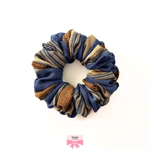 May include: A blue and brown patterned scrunchie with a label that reads "chunky ties". The scrunchie is made of fabric and is circular in shape. The fabric has a denim-like appearance with brown and gold accents.