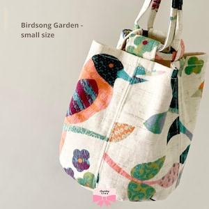 May include: Small tote bag with a bird and floral design. The off-white bag features a bird in orange, teal, and purple, with green leaves and flowers. The text "Birdsong Garden - small size" is visible. The bag has a handle.