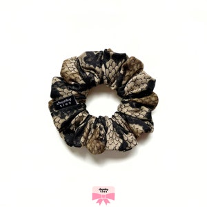 Puede incluir: Una goma para el pelo con un estampado de piel de serpiente en negro, marrón y dorado. La tela está fruncida y plisada, creando una apariencia texturizada. Una pequeña etiqueta negra con el texto "chunky ties" está adjunta. La goma está diseñada para peinar el cabello.