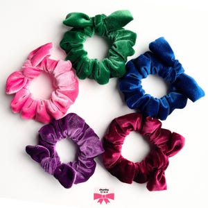 May include: A collection of six velvet scrunchies in various colors: pink, green, blue, purple, and burgundy. Each scrunchie is made of soft velvet fabric and features a bow detail. The scrunchies are arranged on a white surface.
