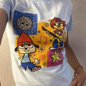 May include: A light blue short-sleeve t-shirt featuring a colorful graphic print. The design includes a cartoon character in a red hat, a character playing a yellow guitar, a moon in a blue starry square, a yellow star, and musical notes.