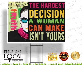 May include: A black and white image of Ruth Bader Ginsburg with the text "The Hardest Decision A Woman Can Make Isn't Yours". The text is in rainbow colors with a distressed look. The image is on a yard sign with the words "Feels Like Local" at the bottom.