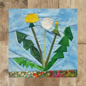 Könnte beinhalten: Ein blauer Stoff-Patchworkblock mit einem Design aus drei weißen Löwenzahnpflanzen mit grünen Blättern und Stielen. Die Löwenzahnpflanzen haben weiße Samen, die im Wind fliegen. Der Patchworkblock hat einen Rand aus kleinen Blumen in verschiedenen Farben.