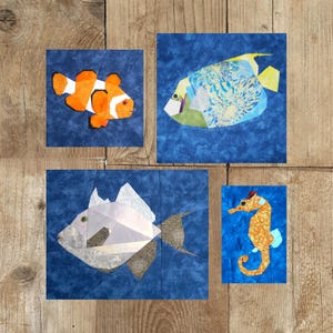 Könnte beinhalten: Vier Stoffkunstquadrate mit Meeresbewohnern auf blauem Hintergrund. Ein Clownfisch mit orange-weißen Markierungen, ein Fisch mit blau-gelben Mustern, ein silberner Fisch und ein orangefarbenes Seepferdchen mit blauen Akzenten.