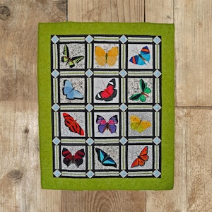May include: A green fabric wall hanging with a grid of 12 squares. Each square features a different butterfly design with various colours, including red, blue, yellow, green, and black.