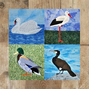 Op de afbeelding: Een patchworkdeken met vier verschillende vogels: een witte zwaan, een zwart-witte ooievaar, een groene eend en een bruine aalscholver. Elke vogel is geplaatst tegen een blauwe of groene achtergrond.