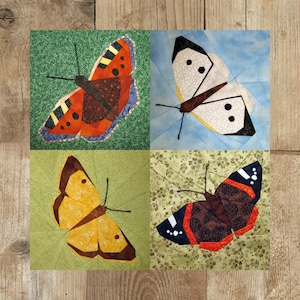 Op de afbeelding: Een quiltblok met vier verschillende vlinderontwerpen. De vlinder linksboven is oranje, zwart en wit. De vlinder rechtsboven is wit met zwarte stippen. De vlinder linksonder is geel met bruine markeringen. De vlinder rechtsonder is zwart, rood en wit.