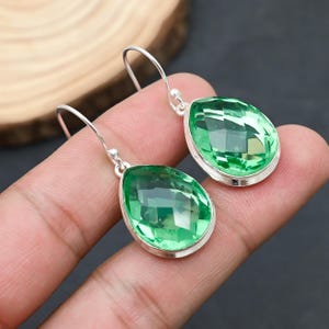May include: A pair of silver drop earrings featuring large, faceted, pear-shaped green gemstones. The gemstones are set in a silver bezel, and the earrings have a simple hook closure. The earrings are displayed against a dark background.