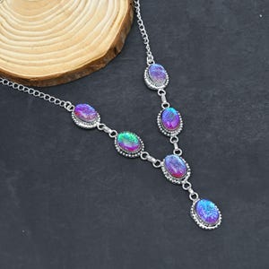 Könnte beinhalten: Eine silberfarbene Halskette mit ovalen, irisierenden Steinen in Lila, Blau und Grün. Die Steine sind in einem dekorativen Silberrahmen gefasst und durch eine filigrane Kette verbunden. Die Halskette wird auf dunklem Untergrund präsentiert.