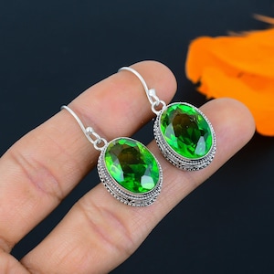 May include: A pair of silver earrings featuring large, oval, bright green gemstones. The gemstones are set in a detailed, ornate silver setting. The earrings are shown against a dark background with a hint of orange.