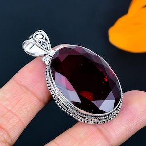 Puede incluir: Colgante de plata con una piedra preciosa ovalada, facetada, de color rojo intenso. El colgante tiene una fianza de plata adornada y un borde decorativo de plata alrededor de la piedra preciosa. La piedra preciosa es el punto focal, captando la luz y mostrando su rico color.