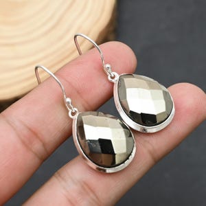 Può includere: Un paio di orecchini pendenti argentati. Ogni orecchino presenta una pietra di pirite sfaccettata a forma di goccia, incastonata in una lunetta d'argento. Gli orecchini hanno una semplice chiusura a gancio.