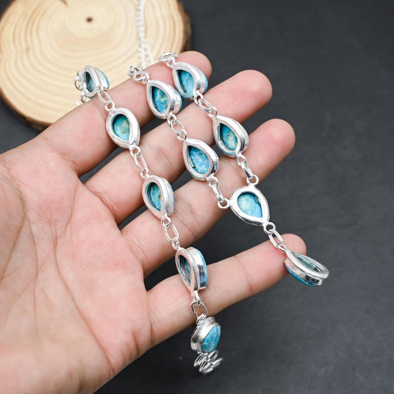Peut inclure: Un collier en argent orn&eacute; de pierres turquoise en forme de goutte d'eau. Chaque pierre est sertie dans un cadre argent&eacute; et reli&eacute;e par de petits maillons. Le collier est pr&eacute;sent&eacute; sur un fond sombre.