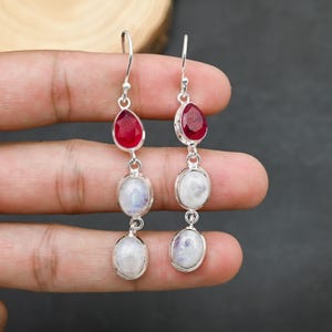 May include: A pair of dangling earrings featuring a teardrop-shaped red gemstone at the top, followed by two oval-shaped, white gemstones. The earrings are connected by silver metal links.