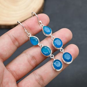 May include: A pair of long, dangling earrings with a silver metal setting. Each earring features three oval and teardrop-shaped, faceted blue gemstones. The earrings are displayed against a dark background.