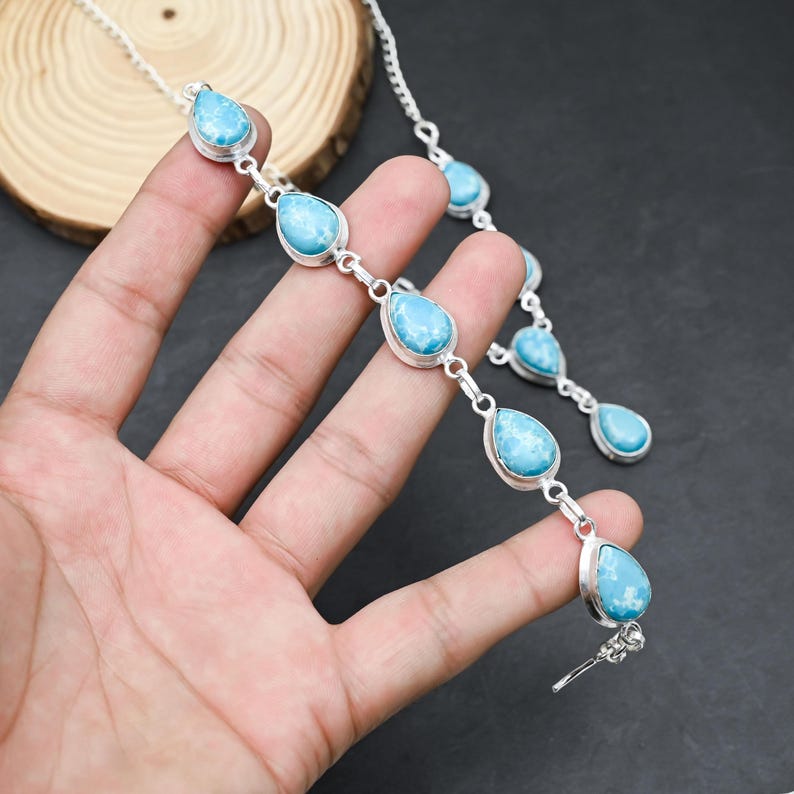Peut inclure: Un ensemble collier et bracelet en argent orn&eacute; de pierres bleues claires en forme de goutte. Les pierres sont serties dans des montures en argent et reli&eacute;es par des cha&icirc;nes en argent. Les bijoux sont pr&eacute;sent&eacute;s sur une surface sombre.