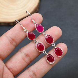 May include: Dangle earrings with a silver-tone metal setting. Each earring features three red oval and teardrop-shaped gemstones. The top gemstone is a deep pink color. The earrings are displayed against a dark background.