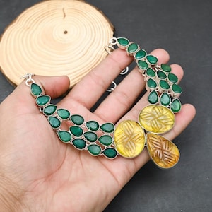 May include: A silver necklace featuring green teardrop-shaped stones and yellow and brown carved stones. The necklace is displayed on a hand against a dark background. The necklace has a toggle clasp.