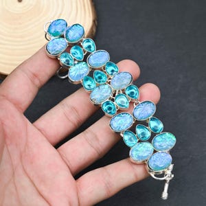 Op de afbeelding: Een zilveren armband met ovale en druppelvormige blauwe en turquoise edelstenen. De edelstenen zijn gezet in een zilveren metalen frame. De armband heeft een veilige sluiting en is ontworpen om om de pols te worden gedragen.