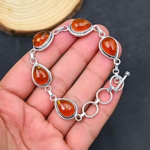 May include: A silver bracelet featuring teardrop-shaped amber stones. The amber stones are a warm orange color and are set in a silver bezel. The bracelet has a toggle clasp and is displayed on a hand.