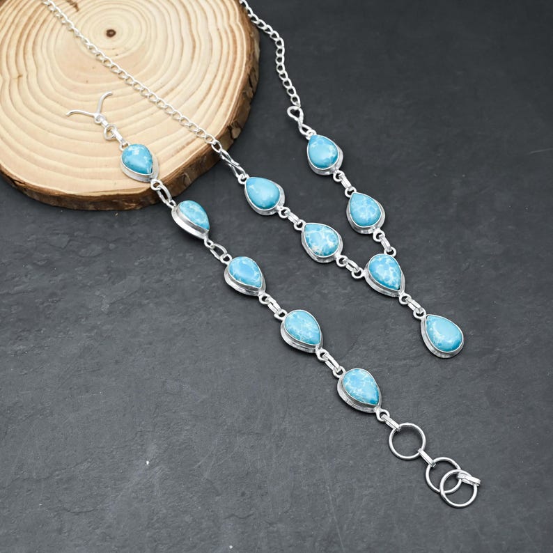 Peut inclure: Un ensemble collier et bracelet en argent orn&eacute; de pierres pr&eacute;cieuses bleues claires en forme de goutte. Les bijoux sont pr&eacute;sent&eacute;s sur une surface en bois sur fond sombre. Le collier a une cha&icirc;ne et le bracelet un fermoir.