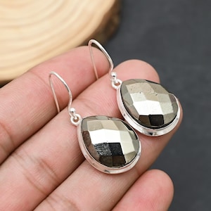 May include: A pair of silver drop earrings featuring faceted, pear-shaped pyrite stones. The pyrite stones have a metallic, reflective appearance, set in a simple silver bezel. The earrings are designed with a hook closure.