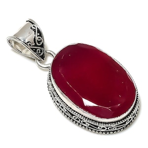 Könnte beinhalten: Ein silberner Anhänger mit einem ovalen, tief roten Edelstein, der in einen silbernen Rahmen mit filigranen Designs gefasst ist.