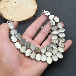May include: A multi-strand statement necklace featuring numerous oval-shaped metallic pyrite stones set in silver-toned metal bezels. The stones exhibit a reflective, textured surface with varying shades of grey, gold, and silver, arranged in a curved, layered design.