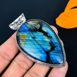 Puede incluir: Un colgante en forma de lágrima con una llamativa piedra de labradorita azul y amarilla. La piedra está engastada en un metal plateado con una fianza ornamentada. El colgante se sostiene en una mano sobre un fondo oscuro.