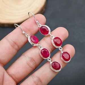 May include: Silver dangle earrings featuring three oval-cut, vibrant red gemstones. The gemstones are set in a silver bezel setting and suspended from a silver hook. The earrings are displayed against a dark background.