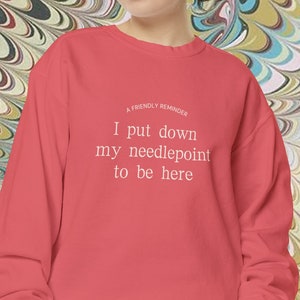 May include: A red sweatshirt with the text "A friendly reminder I put down my needlepoint to be here" printed in white.