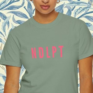 May include: A sage green t-shirt with the word "NDLPT" printed in hot pink.