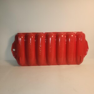 May include: A bright red, vintage-style hot dog bun baking pan. The pan has seven individual molds, each shaped like a hot dog bun. The pan has a rectangular shape with rounded edges and small handles on each end.