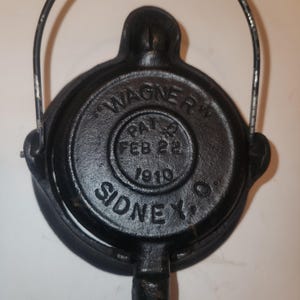 May include: A black cast iron waffle iron with a handle and a hinged design. The iron is marked with the text "WAGNER", "PAT. FEB 22 1910", and "SIDNEY, O."