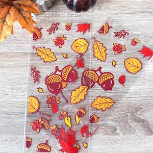 May include: Three clear plastic sheets with autumn-themed designs. The designs include acorns, leaves, and berries in shades of red, orange, and yellow.