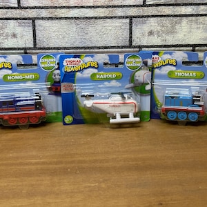May include: Four Thomas & Friends toy train engine packaging boxes. The boxes feature a blue and red train engine, a white helicopter, a blue train engine, and a blue train engine with a yellow face. The boxes have the words "Thomas & Friends Adventures" and "Explore & Imagine" printed on them.