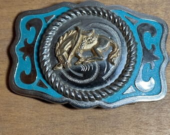 May include: A silver belt buckle with a turquoise blue enamel background. The buckle features a gold-coloured image of a cowboy riding a bucking horse inside a rope circle.