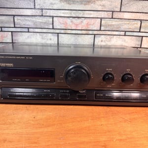 May include: A dark gray Technics stereo integrated amplifier model SU-G91 featuring a large central volume knob, smaller adjustment dials for balance, bass, and treble, a digital display, and various push-button controls including Dolby Surround Pro Logic.