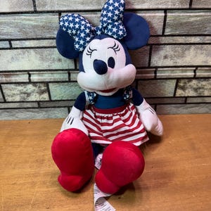 May include: A Minnie Mouse plush doll with a blue bow featuring white stars, blue ears, and a red and white striped skirt. The doll has red shoes and white gloves. The doll is sitting on a wooden surface.